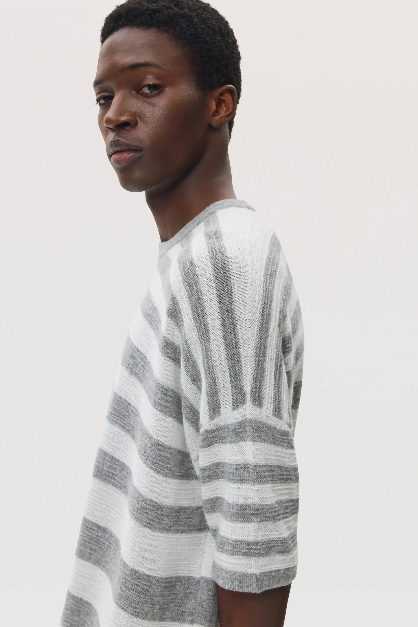 Side view of a male model wearing Johnstons of Elgin SS25 Unisex Knitwear Light Grey & White Textured Stripe Oversized Cashmere T-Shirt on a white backdrop KAA05414Q25124