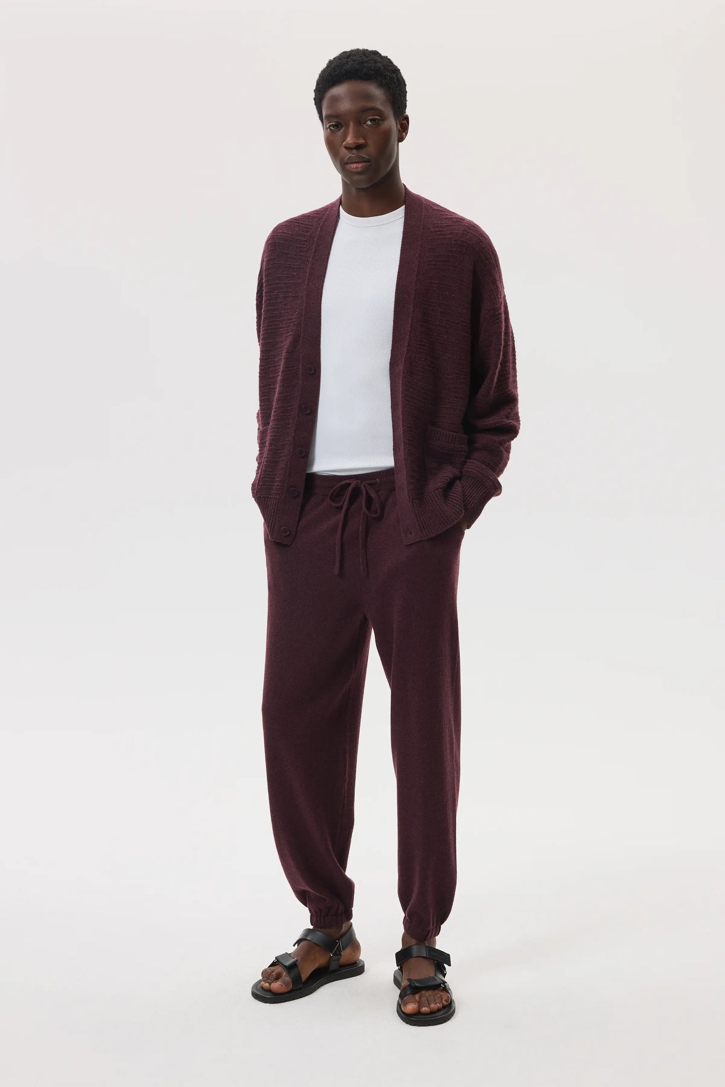 Male model wearing Johnstons of Elgin SS25 Unisex Knitwear Rosewood Textured Cashmere Cardigan with superfine cashmere sweater in white, and rosewood cashmere jogger against a white background KAC05415SB7089