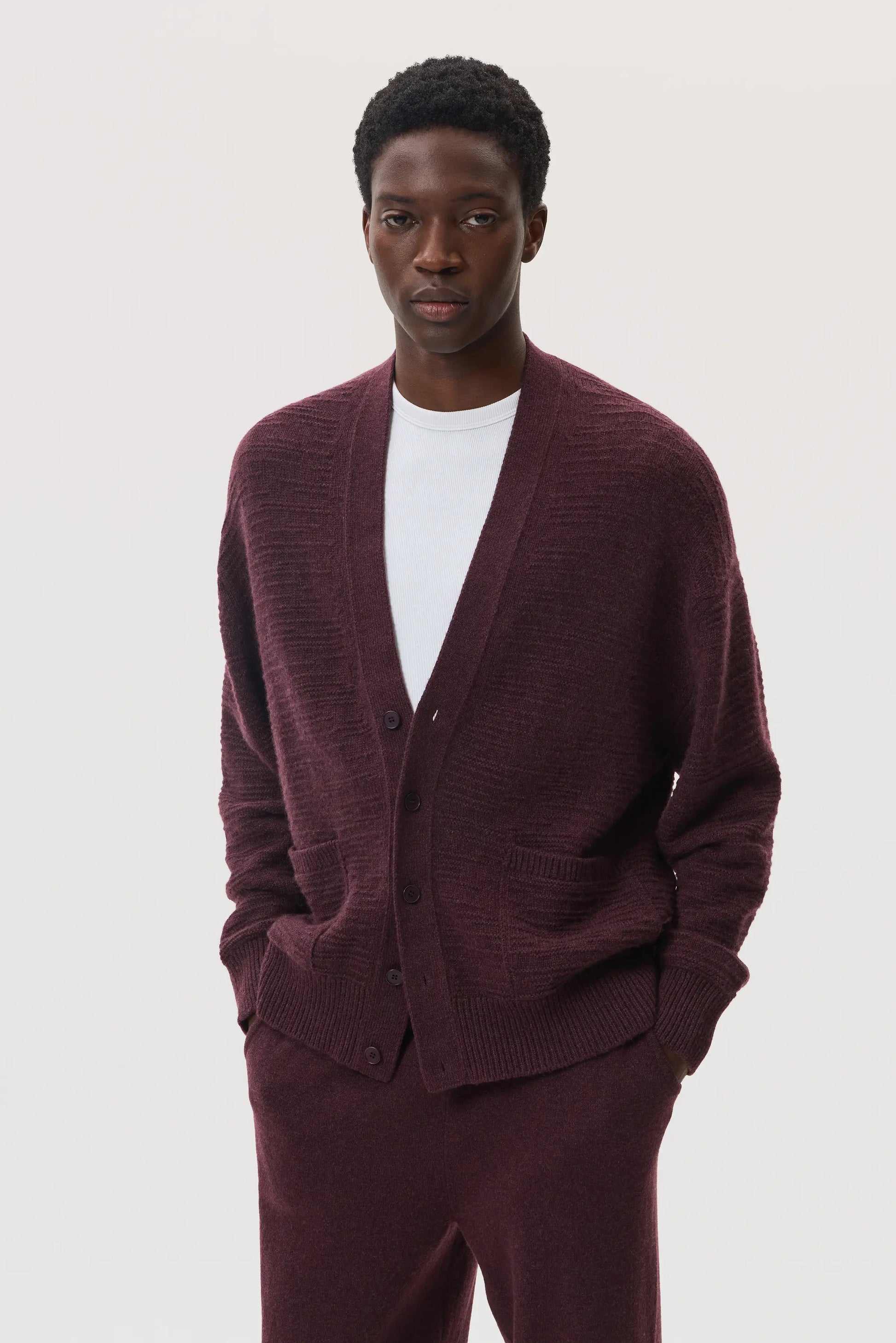 Male model wearing Johnstons of Elgin SS25 Unisex Knitwear Rosewood Textured Cashmere Cardigan with superfine cashmere sweater in white, and rosewood cashmere jogger  KAC05415SB7089