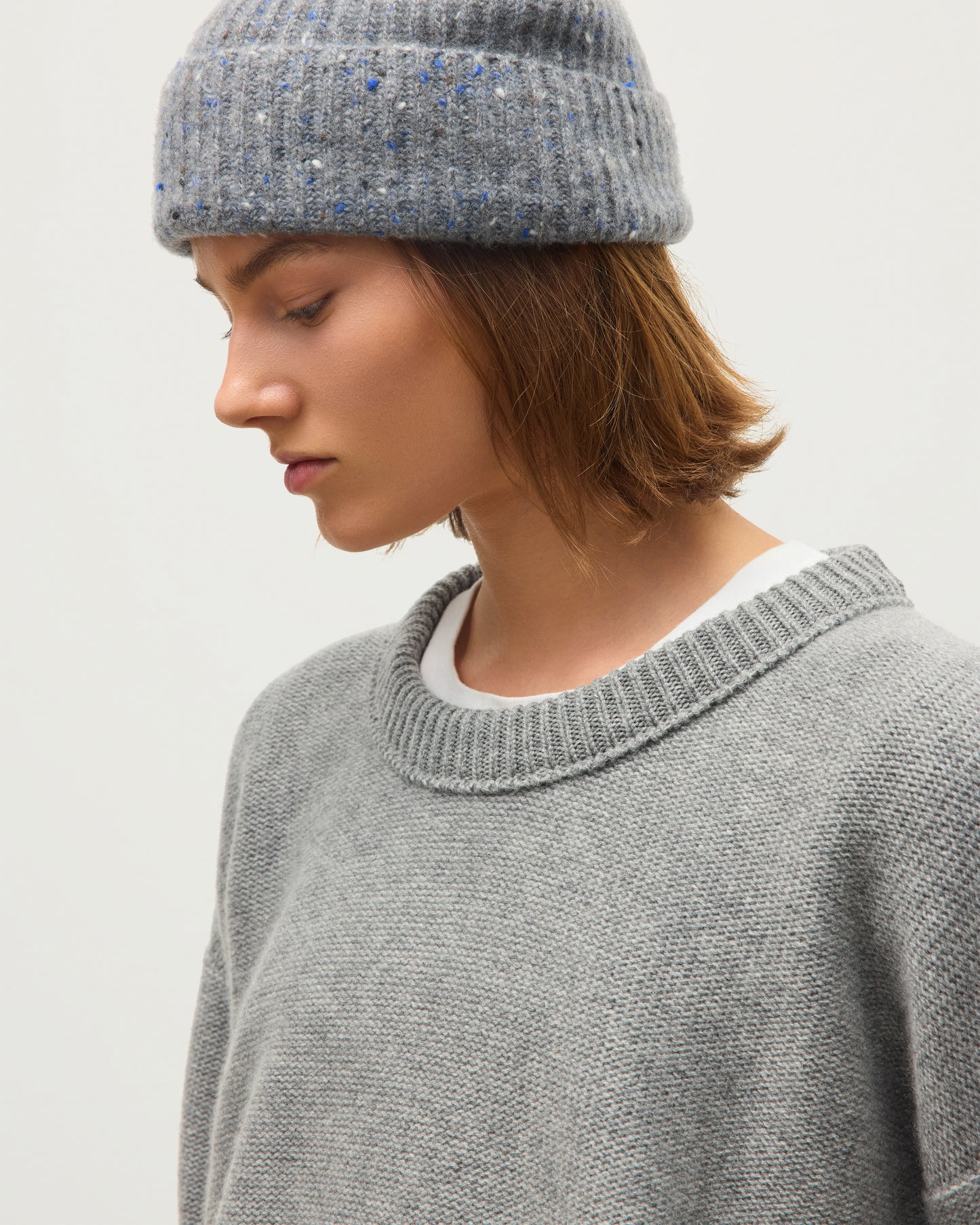 A close-up of a person wearing a gray knit crewneck sweater and a speckled gray beanie against a plain light background. JOHNSTONS OF ELGIN MAGGIE CREW SWEATER CASHMERE REVERSE STITCH ROUND NECK SWEATER - 8 PLY GREY 100% CASHMERE KAI05990HA0400