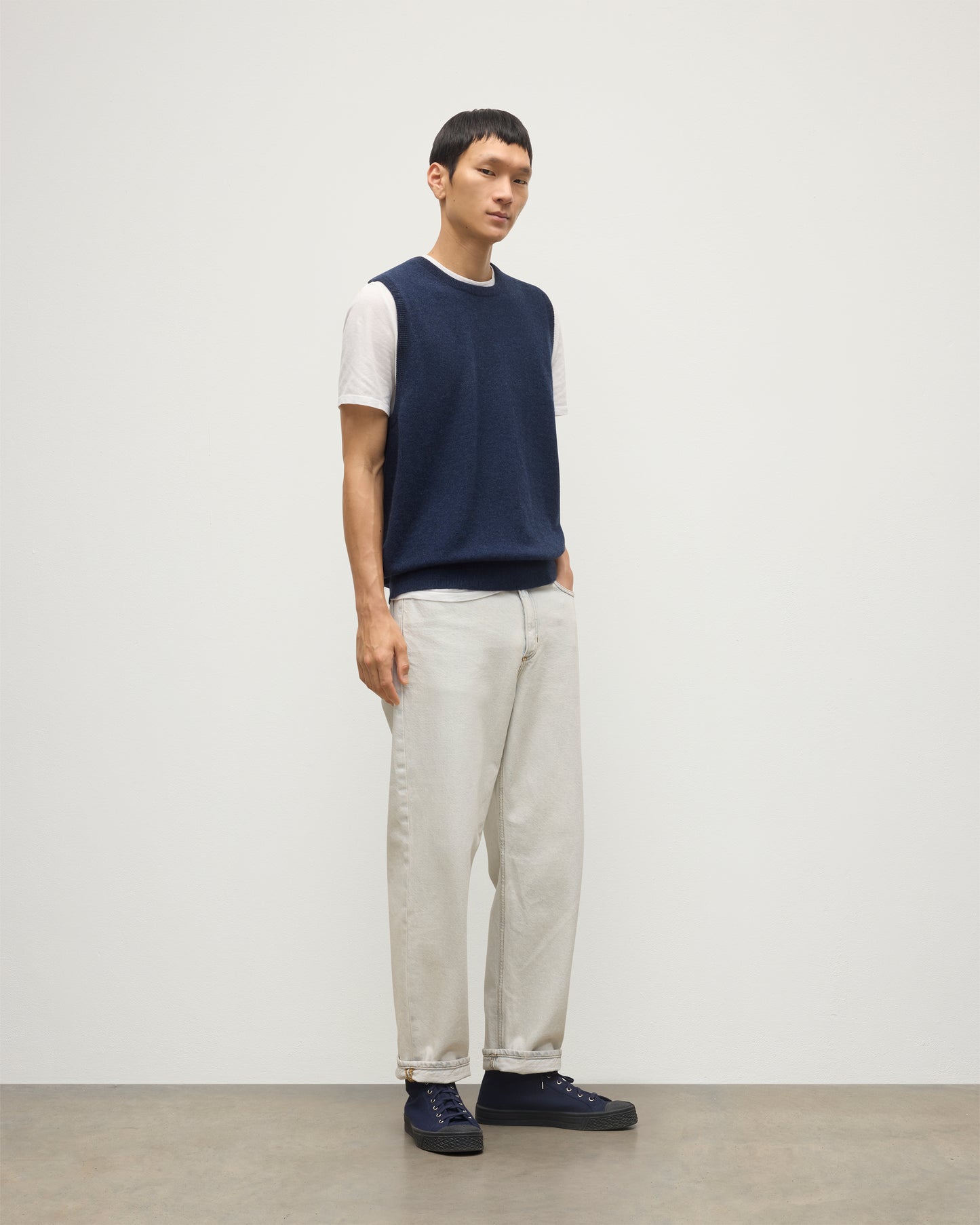 MODEL WEARS JOHNSTONS OF ELGIN MENS LENNOX SLEEVELESS CREW NAVY 100% CASHMERE, OVER A WHITE SHORT SLEEVE T-SHIRT WITH WHITE JEANS KAP05243HD7350
