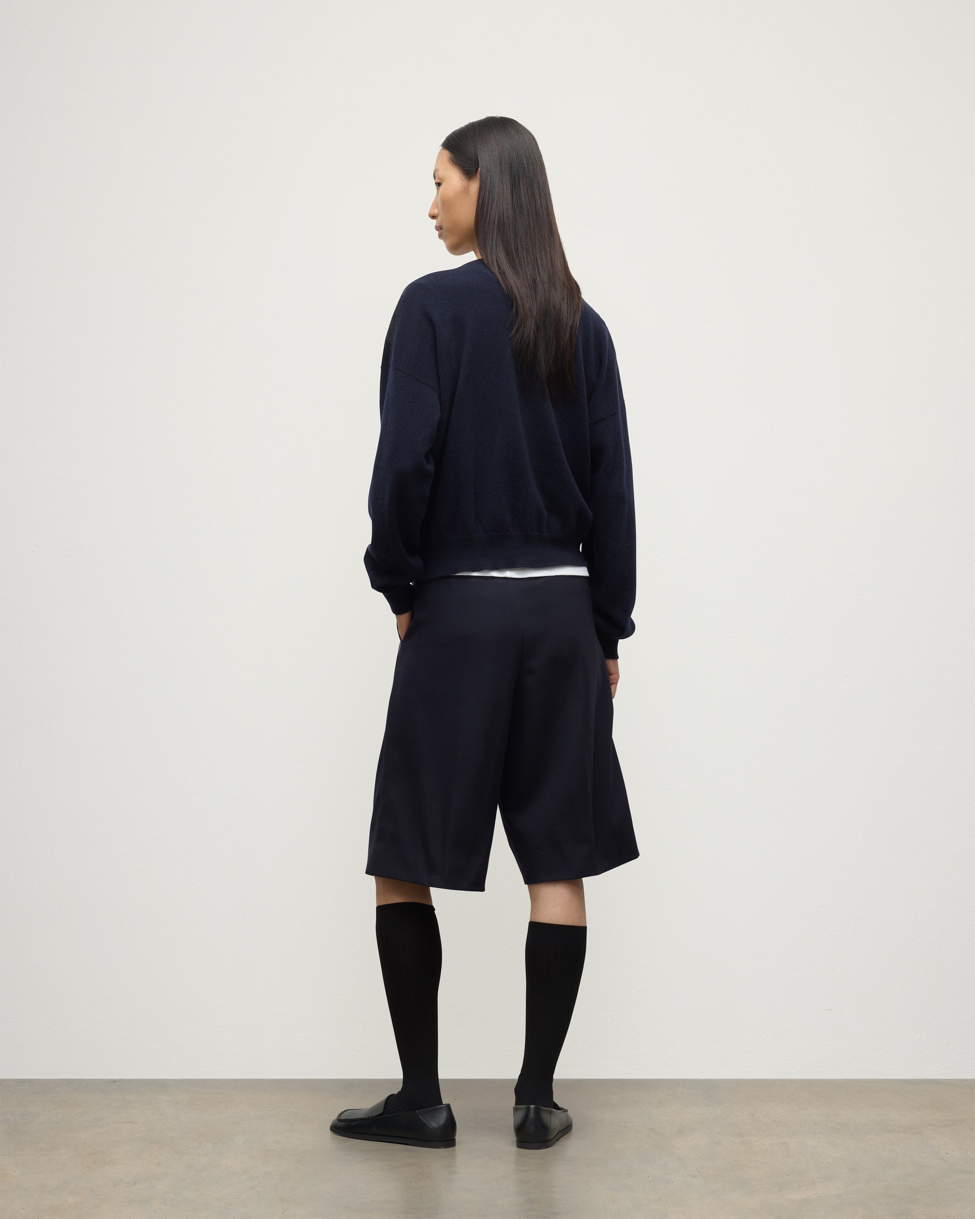 MODEL WEARS JOHNSTONS OF ELGIN WOMENS CLAN SLOUCH CREW NAVY 100% CASHMERE OVER A WHITE TEE, WITH NAVY LONG SHORTS, CASHMERE SOCKS AND LOAFERS KAP05732JU1606
