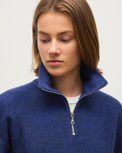 A close-up of a woman wearing a textured blue half-zip sweater with a high collar, layered over a white undershirt, against a plain light gray background. JOHNSTONS OF ELGIN REEL ZIP SWEATER CASHMERE TWILL STITCH ZIP NECK SWEATER - 2 PLY GREY & BLUE 100% CASHMERE KAP06030Q26336
