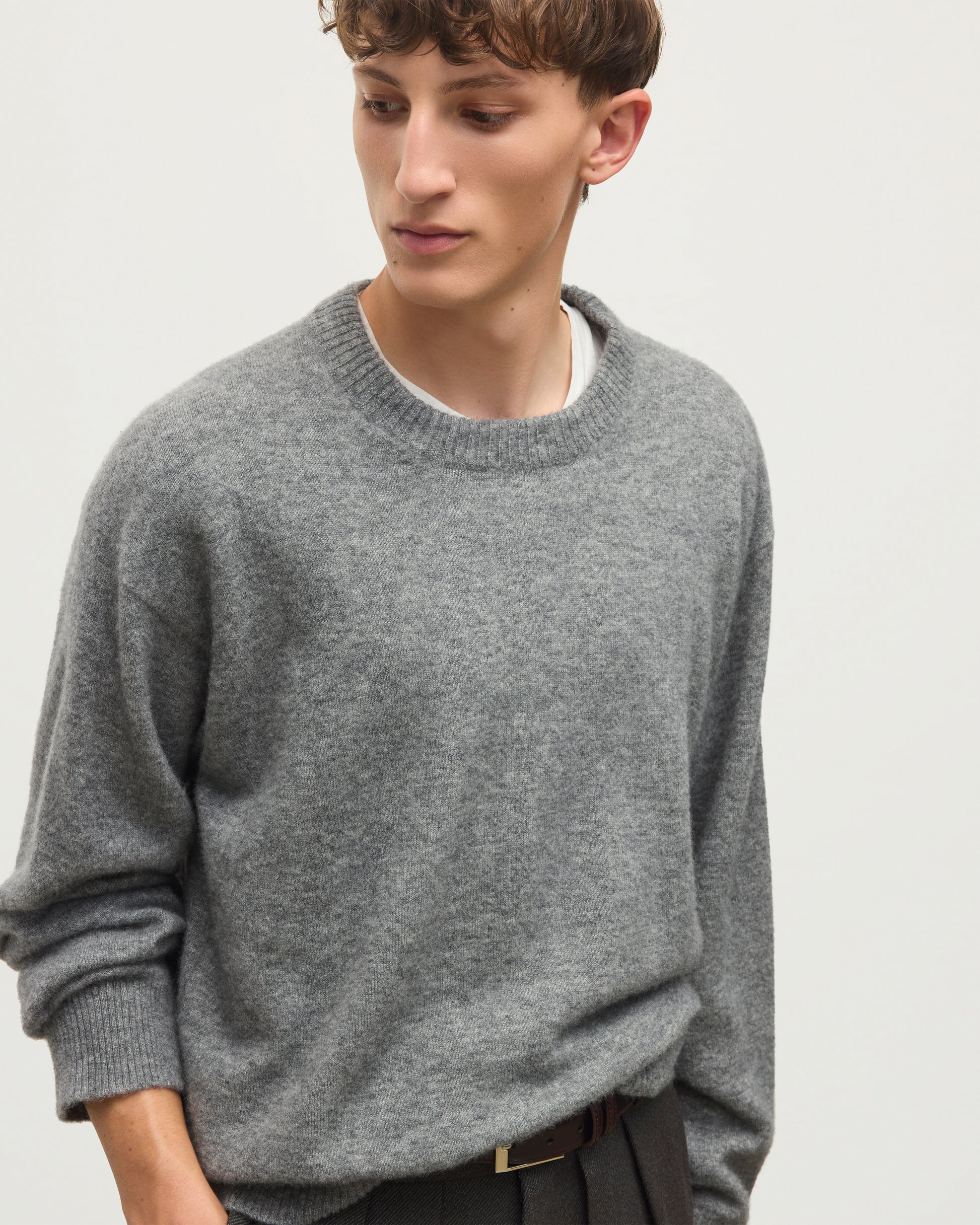 A man wearing a heather gray crewneck sweater over a white undershirt, paired with dark trousers and a brown belt, photographed against a plain light gray background. JOHNSTONS OF ELGIN REUBEN CREW CASHMERE CREW NECK SWEATER - 2 PLY GREY 100% CASHMERE KAP06051SA7216