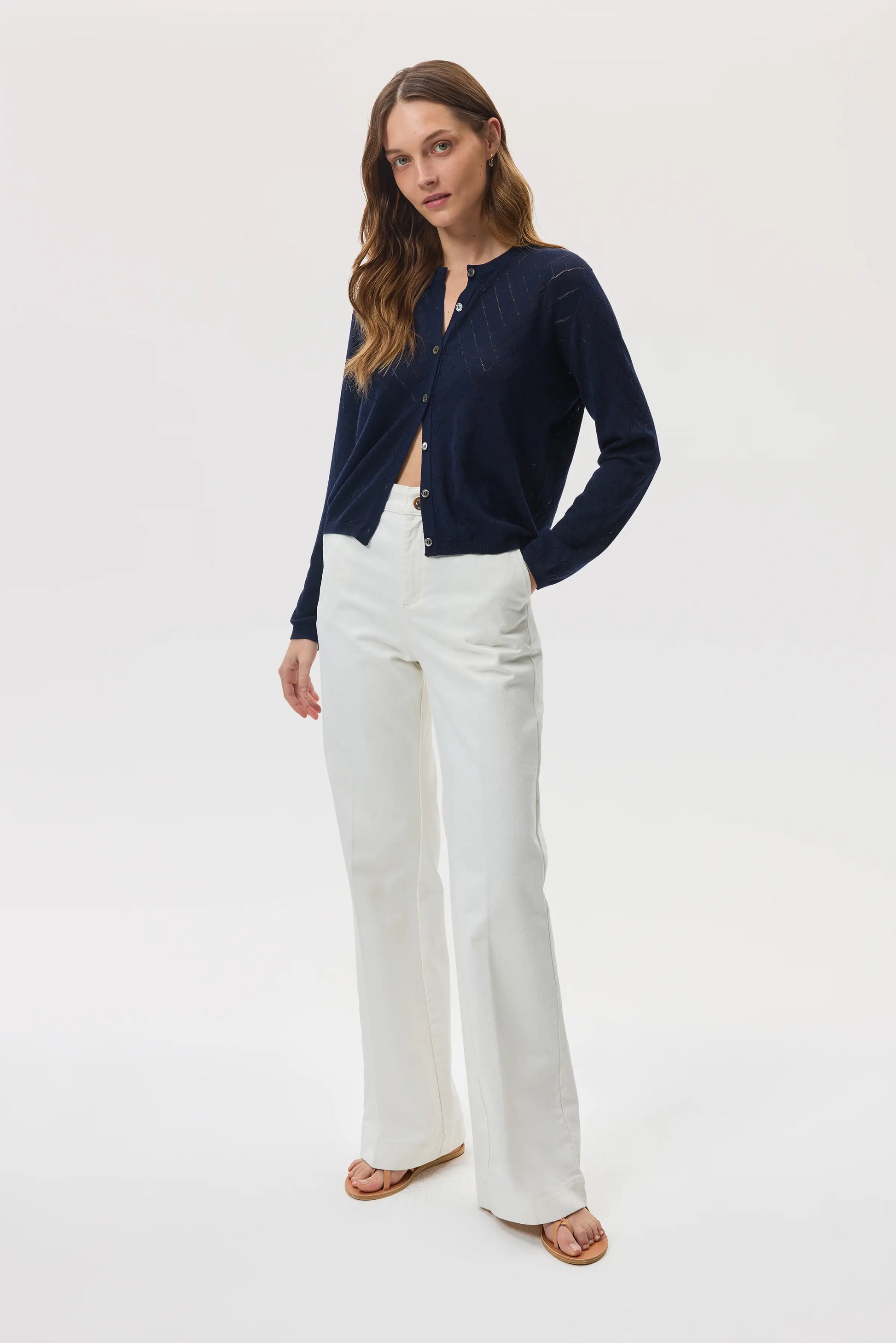 Model wears Johnstons of Elgin SS25 Women's Knitwear Navy Superfine Merino Pointelle Argyle Cardigan with white trousers and sandals KDI00742SD7563