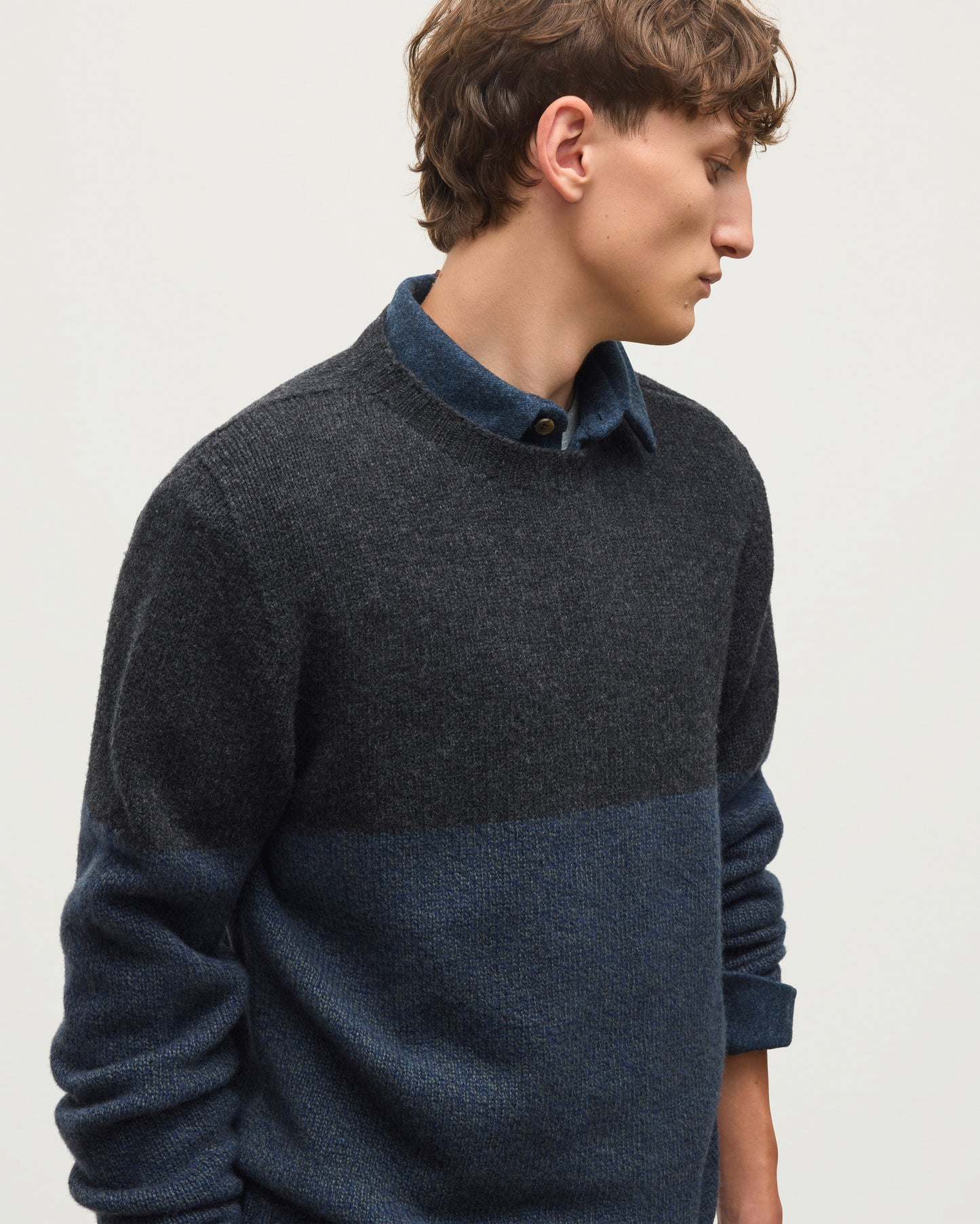 JOHNSTONS OF ELGIN OGILVIE CREW SWEATER LAMBSWOOL COLOURBLOCK CREW NECK SWEATER DARK GREY 100% LAMBSWOOL A man wearing a dark charcoal and navy blue color-block knit sweater over a matching navy collared shirt, paired with black trousers, against a plain light gray background. KDI00816Q26348 