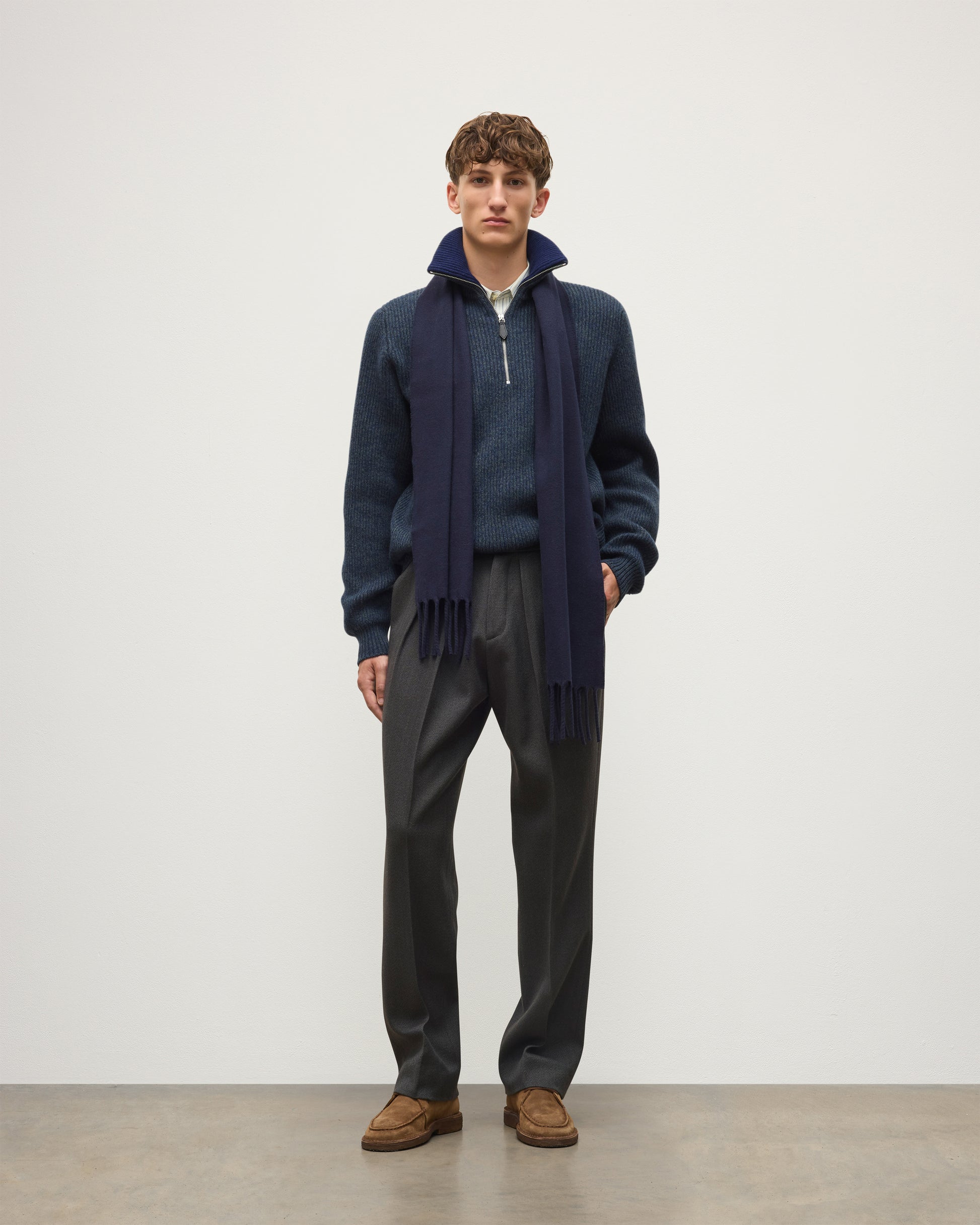 A man stands in a studio wearing a navy quarter-zip sweater over a blue knit top, with a dark navy scarf, dark gray trousers, and brown suede loafers. JOHNSTONS OF ELGIN MACH SCARF DARK NAVY 100% WOOL WD002011SD7330ONE