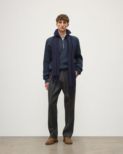 A man stands in a studio wearing a navy quarter-zip sweater over a blue knit top, with a dark navy scarf, dark gray trousers, and brown suede loafers. JOHNSTONS OF ELGIN MACH SCARF DARK NAVY 100% WOOL WD002011SD7330ONE
