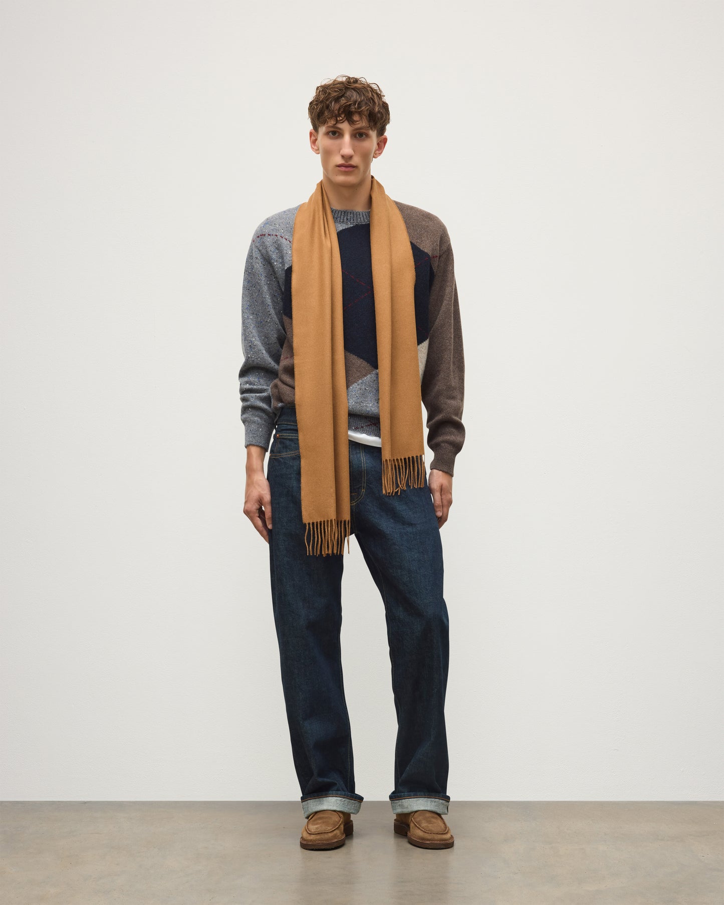 A man stands against a plain light gray studio wall wearing a tan scarf draped over a gray and brown color-block sweater, dark blue jeans, and brown shoes. Johnstons of Elgin Vicuna  Pure Vicuña Scarf WR000025SB3022ONE