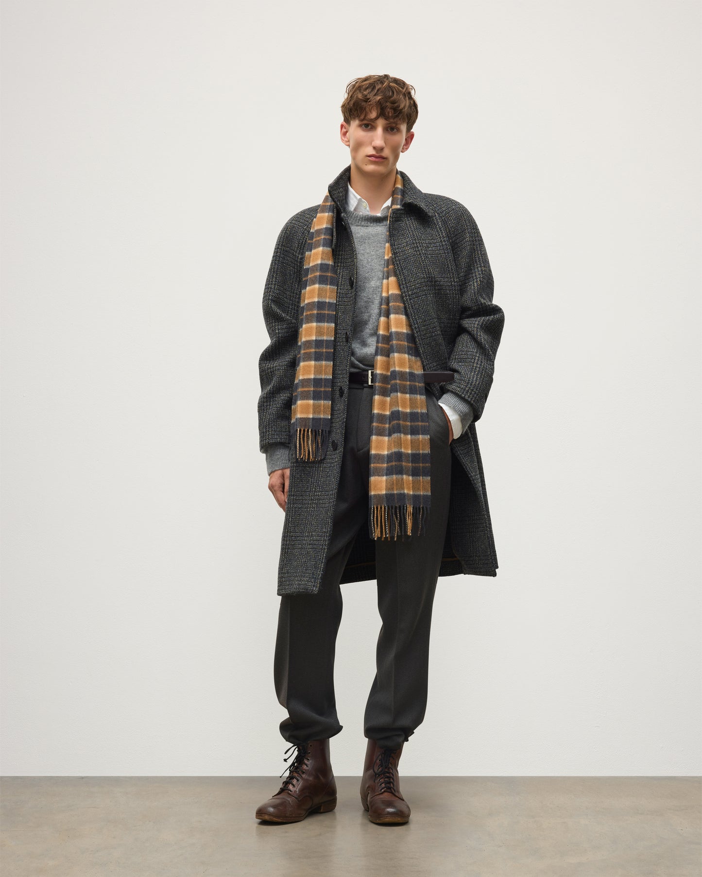 A male model stands against a plain light gray studio background wearing a dark charcoal overcoat, a gray sweater, a brown-and-cream plaid scarf, dark trousers, and brown leather boots. Johnstons of Elgin Vicuna  Windowpane Check Cashmere & Vicuña Scarf WV000013RU6366N/A